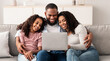 © Prostock-studio - Relaxing Together. Portrait of happy African American parents and their cute daughter using laptop, spending time together, watching movie or surfing internet, sitting on the sofa in living room