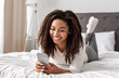 © Prostock-studio - Mobile Phone User. Portrait of happy African American woman lying in bed and smiling while reading text message from someone, holding and using smartphone, chatting with lover or browsing social media