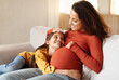 © Prostock-studio - Second Pregnancy, Waiting For Little Brother. Kid girl touching her pregnant mother's belly and smiling to mom at home. Expecting woman sitting with her daughter on couch in living room