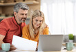 © Prostock-studio - Attractive middle aged spouses working from home, loving husband and wife having family business, sitting at kitchen table in front of laptop, holding documents, embracing, copy space