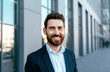 © Prostock-studio - Portrait of smiling confident attractive millennial businessman with beard in suit looking at camera near office building, outdoor, close up. Ceo manager and boss, successful work, career and business