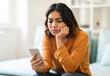 © Prostock-studio - Pensive Young Arab Woman Looking At Smartphone Screen While Sitting On Couch At Home, Upset Millennial Middle Eastern Female Feeling Bored Or Waiting For Important Call Or Sms, Copy Space