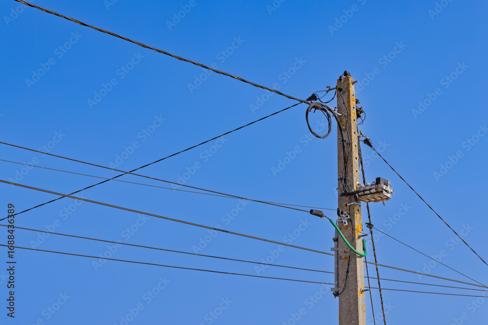A close-up view of a power pole with multiple tangled wires and cables against a clear blue sky. The complex network of lines illustrates urban infrastructure.