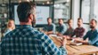 © Green - A bearded man in a blue checkered shirt presents to a diverse group around a table. A chart is visible in the background, suggesting a meeting about analytics