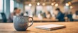 © Shelley - Close-up of a ceramic coffee mug and notebook on a desk in a lively coworking space, with blurred coworkers on laptops in the background. Conveys productivity, focus, and energetic collaboration.