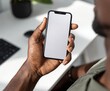 © Basileus - Close-up of a person's hand holding a smartphone with a blank screen, displayed against a backdrop of a workspace.