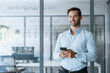 © Stock 4 You - Young European business man manager ceo using cell phone mobile app. Latin Hispanic male businessman holding smartphone in office for trading, reading, working online, smiling at camera. Copy space