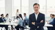 © K99Design - A confident businessman standing in a modern office with his arms crossed, surrounded by colleagues.
