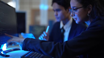 A male and female programmer collaborate on a software project. The team leader is training a new intern, explaining the code on her screen in the tech office after hours.