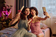 © AnnaStills - Two Caucasian teenage girls sitting on bed holding diplomas and smiling while taking selfie with smartphone, celebrating graduation together in cozy bedroom setting