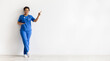 © Anastasiya - African American female nurse in blue scrubs with stethoscope leaning on wall and pointing at copy space. Confident young healthcare professional portrait with white background