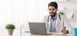 © Prostock-studio - Consult patient online, webinar, conference and meeting at clinic. Friendly young arabian bearded doctor in white coat, glasses with stethoscope look at laptop, makes notes in office interior