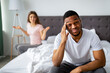 © Prostock-studio - Displeased young guy sitting on bed, massaging temples in irritation, his angry wife shouting at him indoors. Millennial multiracial couple having marriage difficulties, facing relationship problems