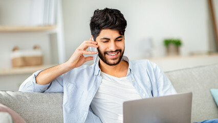 Customer support. Cheerful arab man using laptop talking on phone with call center operator. Freelancer working on computer, sitting on the sofa in living room