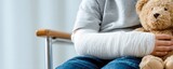 Child with arm cast sits with a teddy bear.  He is wearing a gray shirt and blue jeans and sitting in a chair. Soft, natural lighting
