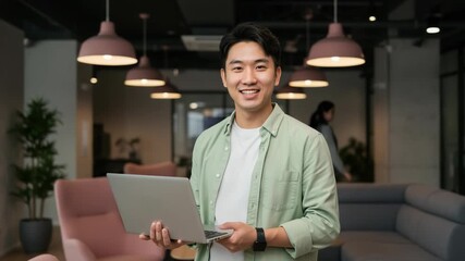 Smiling asian man holding laptop in modern office space