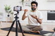 © Sofiia Shunkina - Man sitting cross-legged in kitchen filming a video about healthy eating for viewers. Includes indoor background, white shirt, home setting, casual atmosphere.