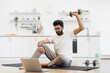 © sofiko14 - Young adult man sits on exercise mat in home kitchen lifting dumbbells by laptop. Engages in remote fitness routine, demonstrating home workout.