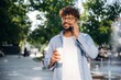 Paper cup in right hand and smartphone in left one. Handsome man is outdoors in the city