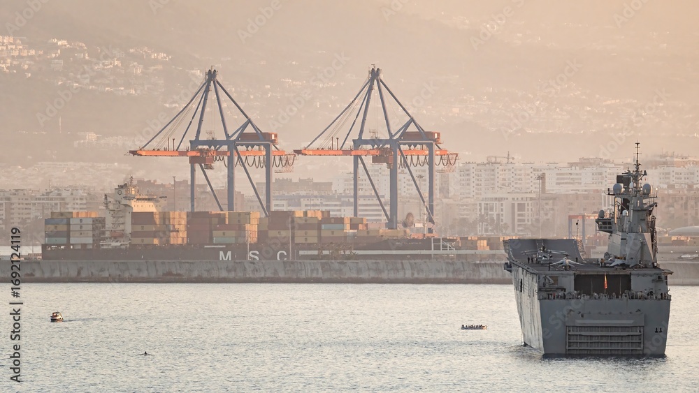 Military ship and container vessel in busy seaport at sunset, with ...