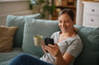 © Stockphotodirectors - A woman smiles as she interacts with her smartphone while holding a cup of coffee. She sits comfortably on a couch in a relaxed living room filled with natural light.