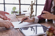 © Tj - Lawyer explaining a legal document to a client, pointing out where to sign, with scales of justice and a gavel prominently displayed on a wooden table in a professional office setting