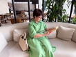 © satura_ - Middle-aged woman in green dress reading book on sofa. Knowledge, leisure, and personal time in modern lifestyle.