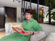 © satura_ - Middle-aged woman in green dress reading book on sofa. Knowledge, leisure, and personal time in modern lifestyle.