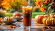 © AnjumanAra - Photo of a refreshing glass of iced pumpkin spice latte sits on a wooden table outdoors, surrounded by mini pumpkins, autumn leaves, and vibrant fall flowers