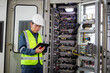 © JD Studio - Inspector wearing safety helmet and high visibility vest is controlling high voltage energy system using tablet in industrial control room with complex wiring and equipment