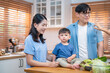 © Thitiporn - Asian parents with their young son in a bright kitchen setting, sitting together at a wooden counter with fresh vegetables, a calm family moment at home.