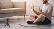 © Prostock-studio - Health care, sports blog, online conference, remote webinar. Happy arabian bearded young 30s guy sits on mat, shows hand to bottle of water in living room interior, looks at webcam of phone on floor