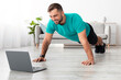 © Prostock-studio - Smiling young guy with beard does plank or push-ups and looks at laptop in living room interior. Body care, workout at home, social distance and device for sports. Training for muscle, online lesson