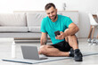 © Prostock-studio - Happy smiling young caucasian guy sitting on mat on floor typing on phone and looking at computer in living room interior. Covid-19 quarantine and video call, communication remotely and trainer online