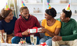 © Prostock-studio - Cute young black friends greeting birthday lady at home, man giving her gift box and smiling. Cheerful african american group sitting on couch, wearing party hats, drinking champagne