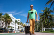 A traveler walks through the sunny streets of Miami. A man traveler enjoys his summer vacation in a tropical city. A traveler tourist with suitcase an urban landscape. Man traveler walk on city.
