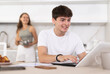 © JackF - Smiling guy doing business on notebook and speaking to his wife standing at the kitchen cabinet