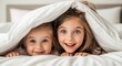 © Artha - Two young girls peek out from under a white duvet, smiling and looking directly at the camera with bright, joyful expressions.