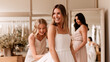 © dinatychynska - Bride trying on simple white wedding dress in bridal salon with friends helping. Happy blonde woman smiling while bridesmaids adjust gown fitting in elegant boutique with natural lighting.