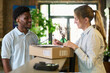 © Mediaphotos - Young Black man delivering package to middle aged woman signing document at counter, both standing indoors with plants in background, engaging in delivery transaction