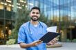 © Liubomir - Portrait of smiling young indian doctor holding medical records outside hospital