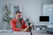 © MINAE - Middle aged male podcaster reading book into microphone while recording podcast episode in home studio, creating educational or entertainment content for online audience