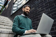 Low angle view of a happy businessman sitting on stairs at downtown district with laptop in his lap and typing on it.
