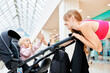 © Georgii - Young pretty woman holding carriage with her child. Happy mother and cute little blonde girl in pink clothes touch hands and laugh. Mom with a baby in a stroller walking around shopping center.