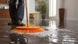 © Oleksandr - Broom cleaning up water on a hard floor. Visible person is wearing brown loafers. In the background you can see a kitchen and living room space.