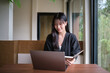© wattana - Young woman works on her laptop while holding a notebook, sitting by a large window with natural light.