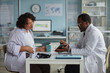 © Seventyfour - Black female patient discussing health concerns with Black middle aged male doctor using digital tablet in medical office, prescription medication and medical equipment on table