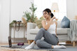 © eakgrungenerd - Asian woman doing online yoga class at home, sitting on yoga mat in butterfly pose, focusing on mindfulness and flexibility, wearing activewear in cozy living room.