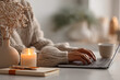 © Jula Isaeva  - Woman typing on laptop near lit candle in cozy home workspace with warm sunlight