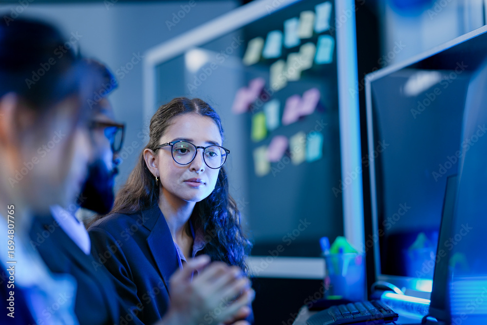 An agile development team participates in a sprint retrospective meeting. A female developer contemplates the user stories on the kanban board visible in the background.